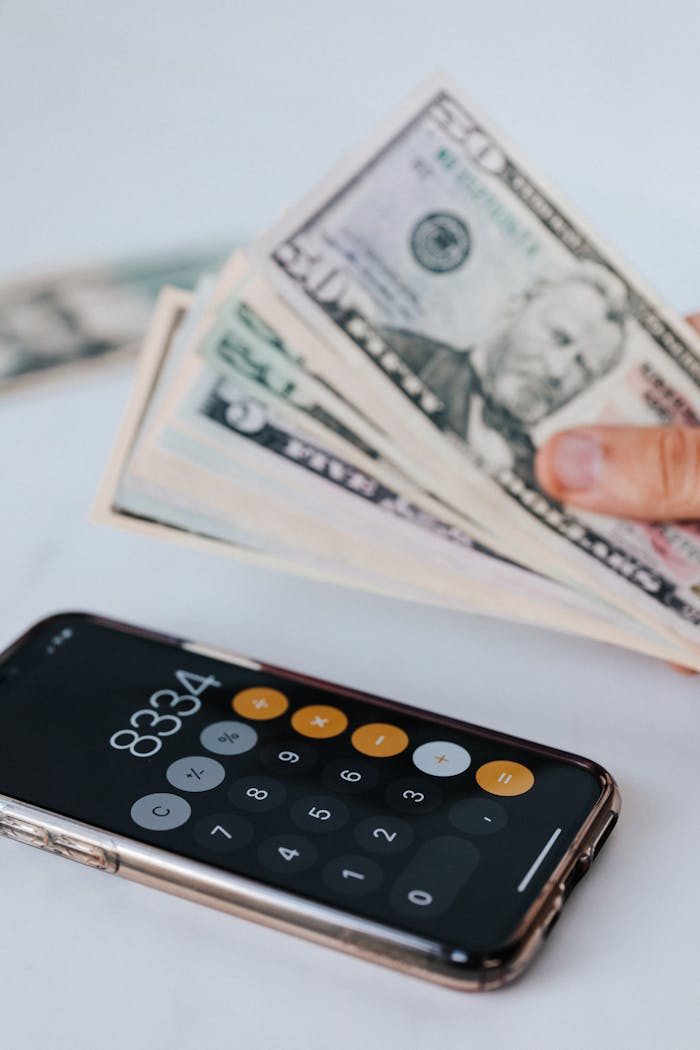 From above of crop anonymous person holding heap of American dollar near cellphone with calculator application representing numbers